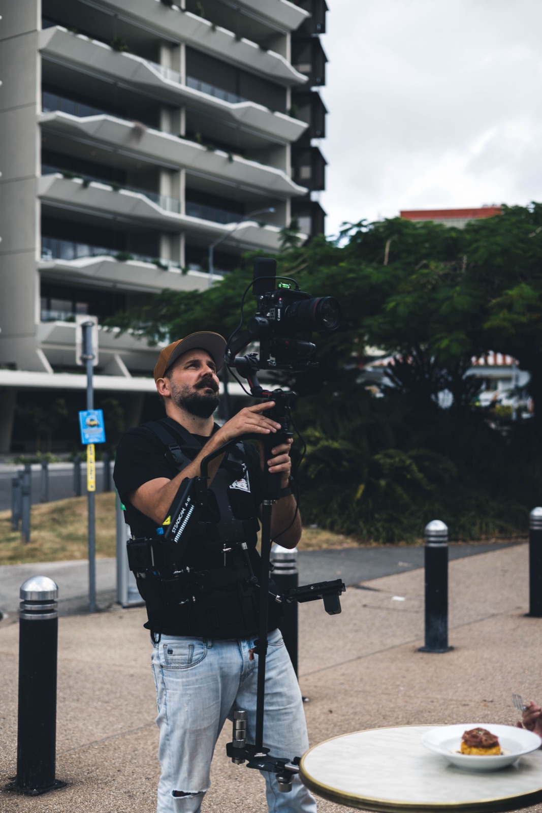 Brisbane content creator operating a Ronin gimbal-stabilised cinema camera against a New Farm high-rise backdrop, dessert plate in foreground.